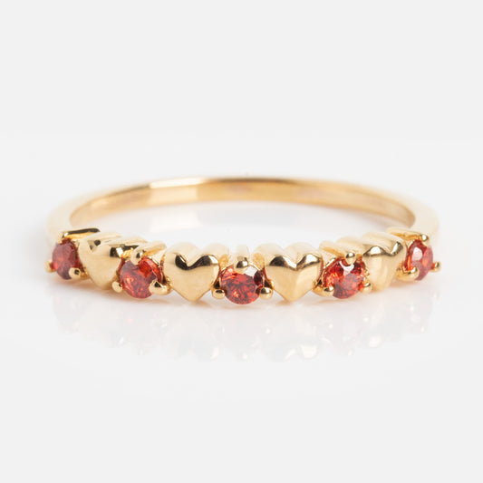 The Sweetheart Stacking Ring is a gold band featuring alternating small heart shapes and round red gemstones arranged in a row, shown against a white background.