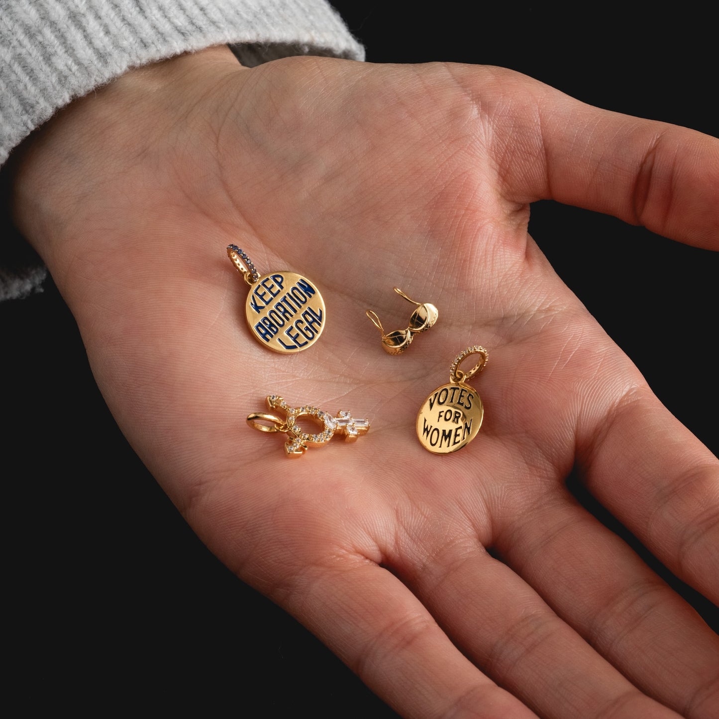 A hand holds three Limited Edition Women's History Month Charms engraved with "KEEP ABORTION LEGAL" and "VOTES FOR WOMEN," along with a pair of gold earrings. The person wears a light, ribbed sweater.
