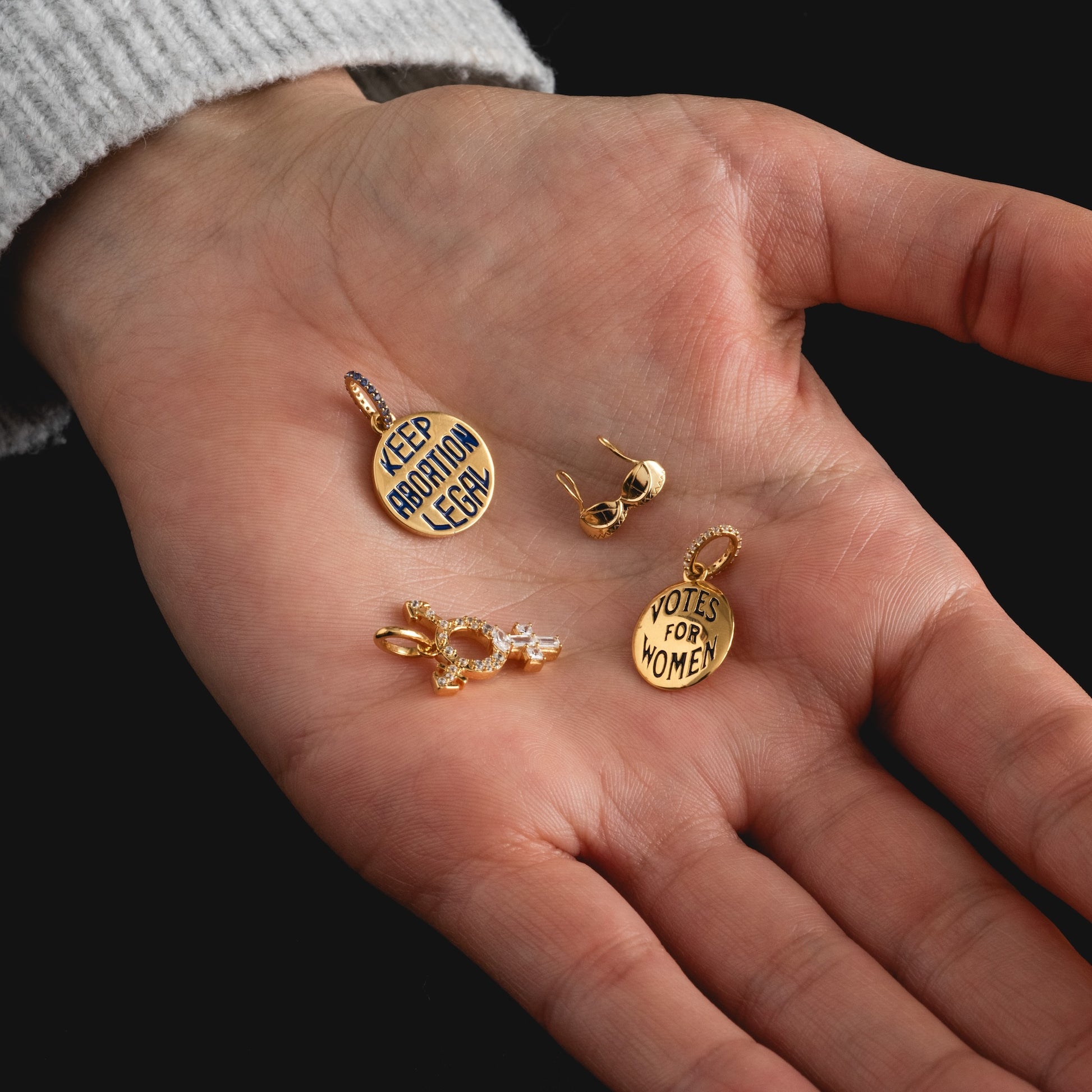 A hand holds three Limited Edition Women's History Month Charms engraved with "KEEP ABORTION LEGAL" and "VOTES FOR WOMEN," along with a pair of gold earrings. The person wears a light, ribbed sweater.