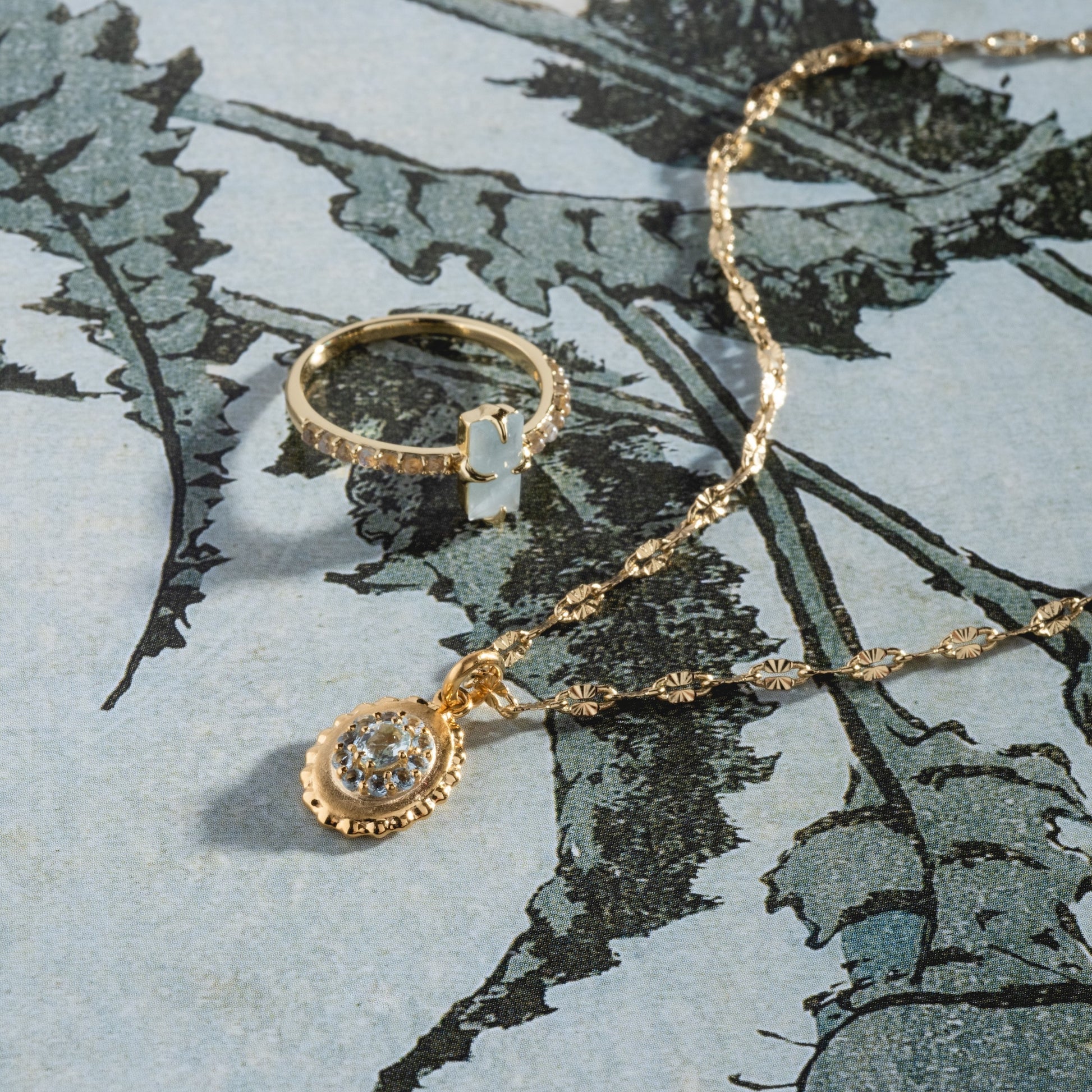 The 2026 March Capsule Vintage Inspired Aquamarine Charm, a gold necklace with a round aquamarine pendant, is displayed on paper featuring a botanical illustration.