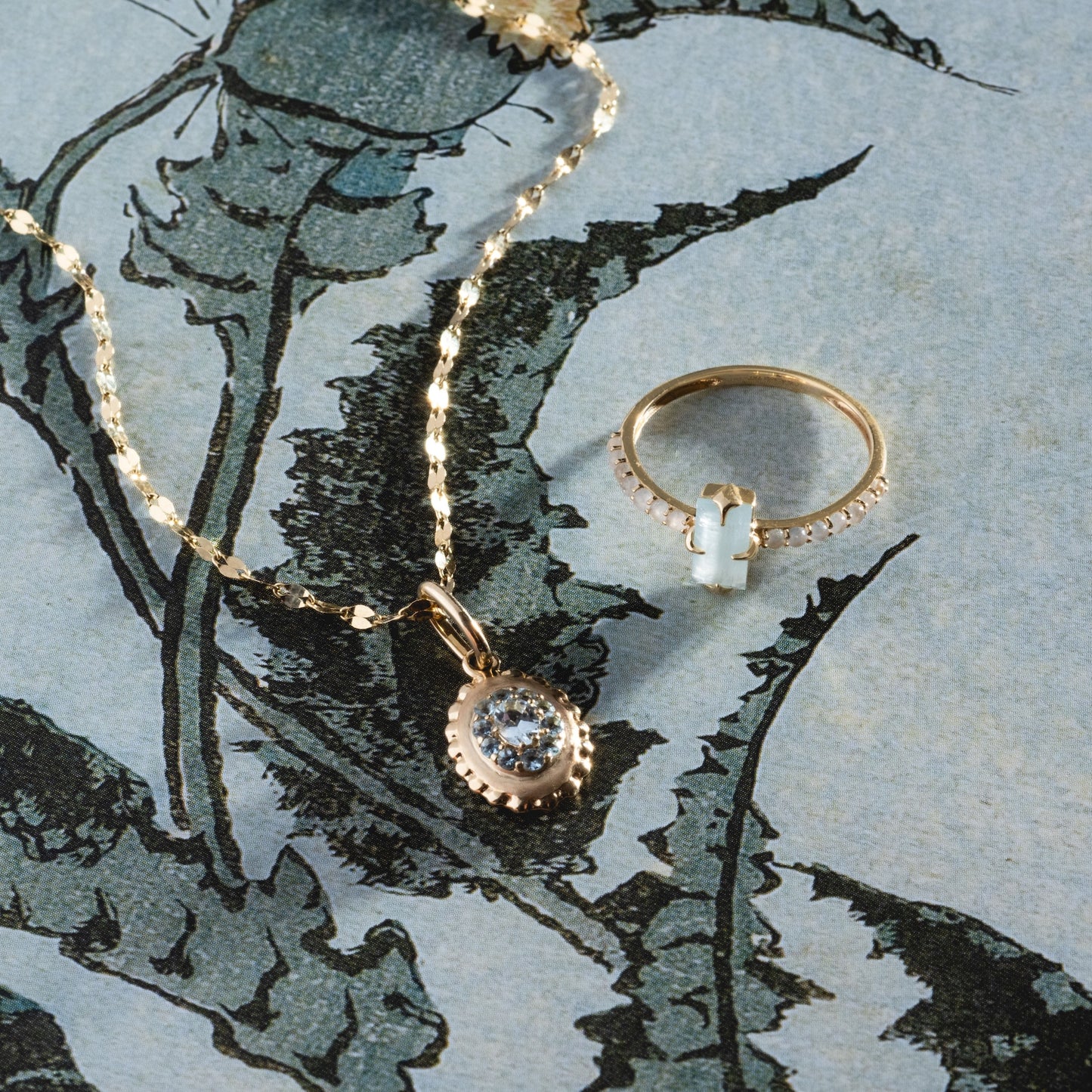 The Solid Gold 2026 March Capsule Vintage Inspired Aquamarine Charm rests on a blue, illustrated floral background alongside a sparkling gold ring with a rectangular gemstone.