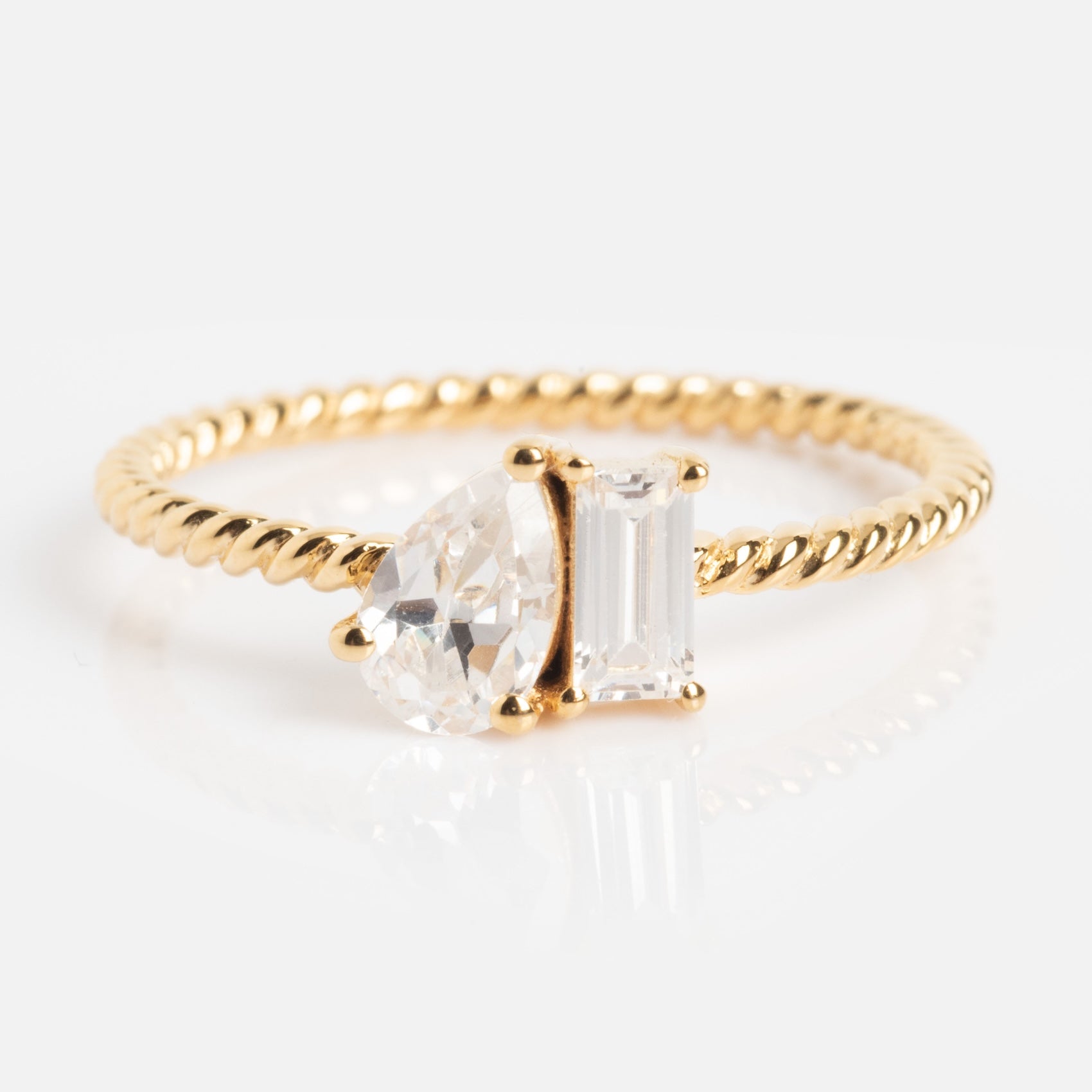 The Twisted Toi Et Moi Ring features a gold twisted band with pear-shaped and emerald-cut clear stones set side by side on a white background.