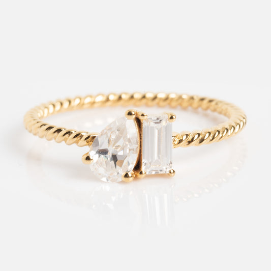 The Twisted Toi Et Moi Ring features a gold twisted band with pear-shaped and emerald-cut clear stones set side by side on a white background.