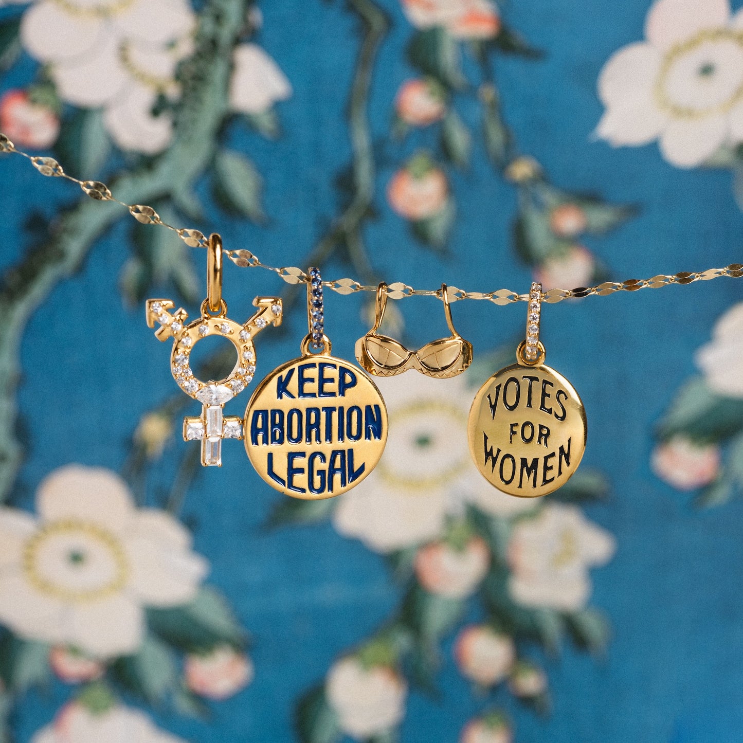 The Limited Edition Women's History Month Charm features four gold chain charms: a rhinestone transgender symbol, a round KEEP ABORTION LEGAL charm, a gold bra, and a VOTES FOR WOMEN charm, shown on a blue floral background.