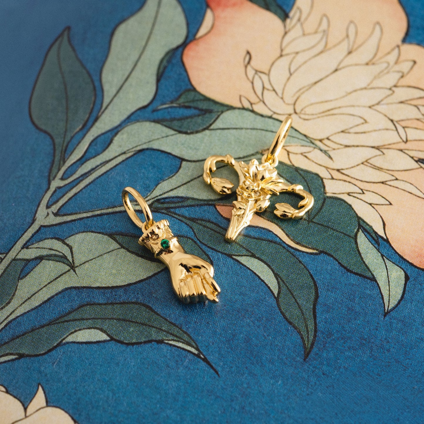 Two 14k yellow gold-plated pendants—a hand with a green CZ gemstone and a ram’s head with curled horns—are featured as the "Free Gift With Orders Over $200 - Choose A Charm." Displayed on a floral-patterned blue background.