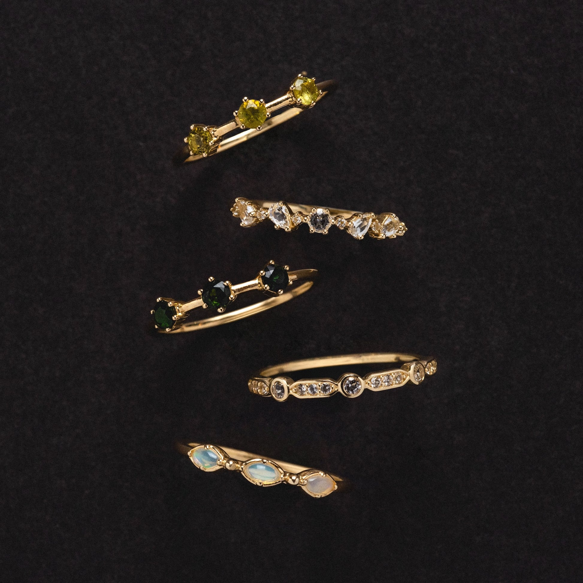 The Solid Gold Tourmaline Trio Stacking Ring, featuring three colorful tourmaline gemstones in an elegant arrangement, is showcased vertically against a dark, textured background.