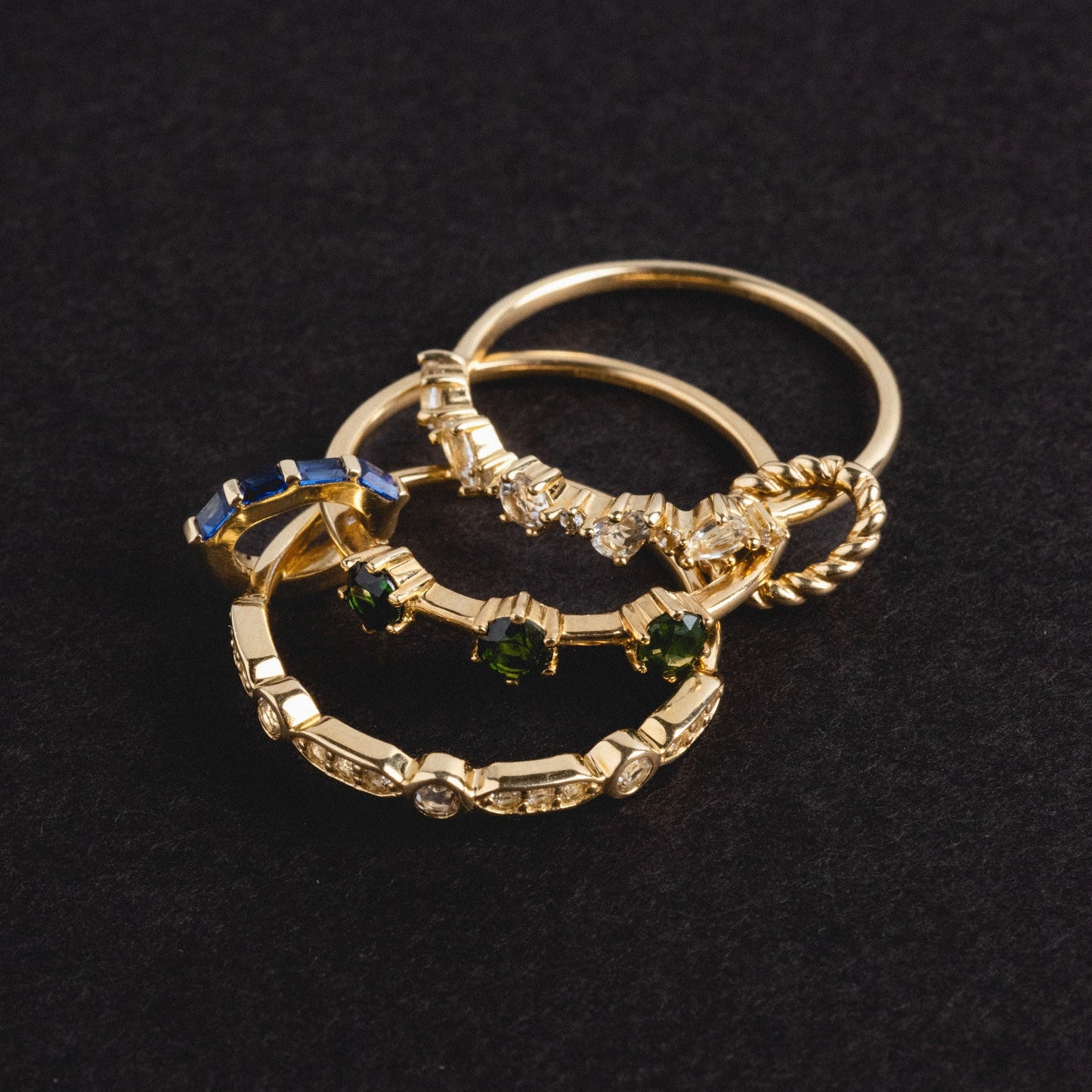 The Solid Gold Tourmaline Trio Stacking Ring, featuring blue and green gemstones in a unique gold design, is displayed overlapping with other gold rings on a black textured background.