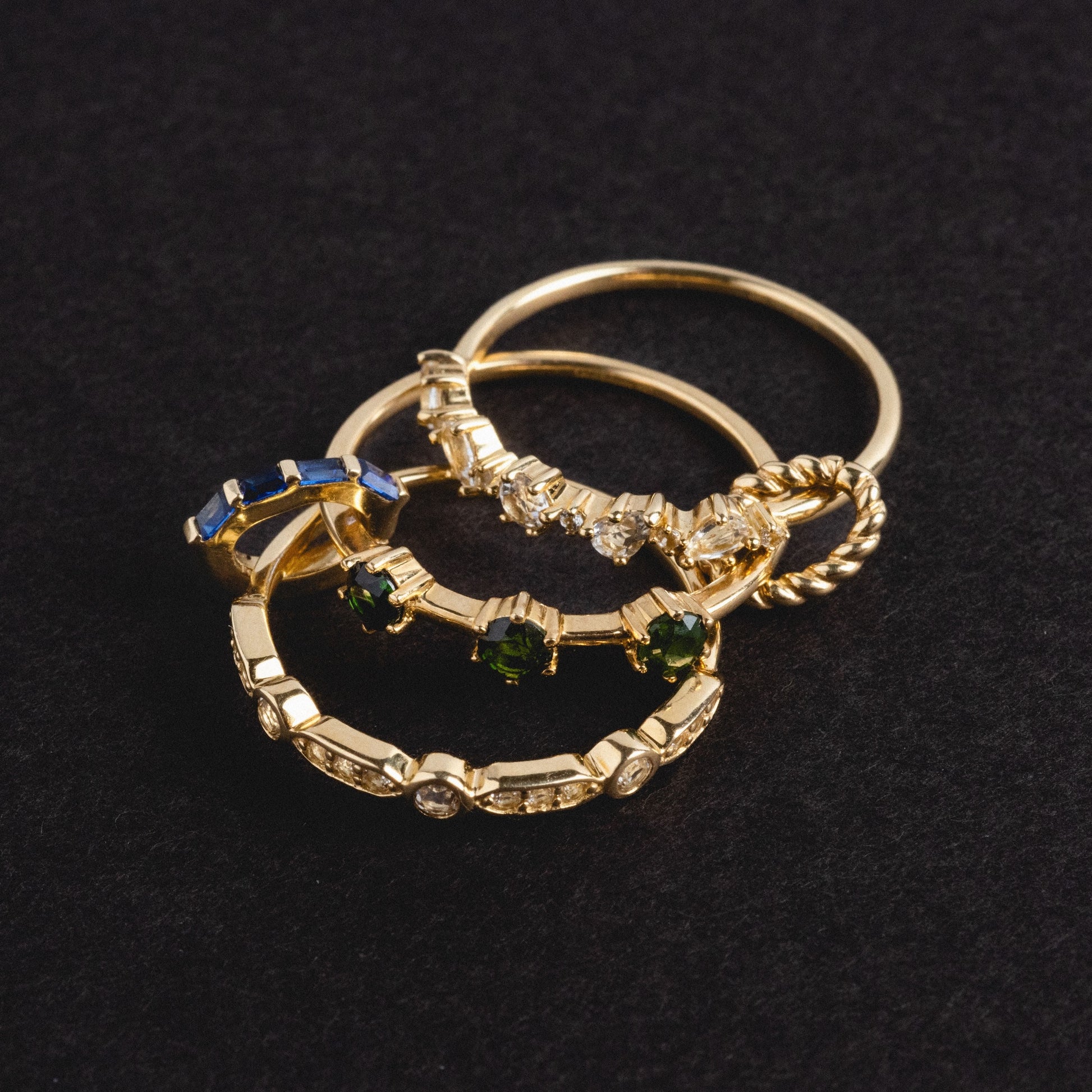 The Solid Gold Tourmaline Trio Stacking Ring, featuring blue and green gemstones in a unique gold design, is displayed overlapping with other gold rings on a black textured background.