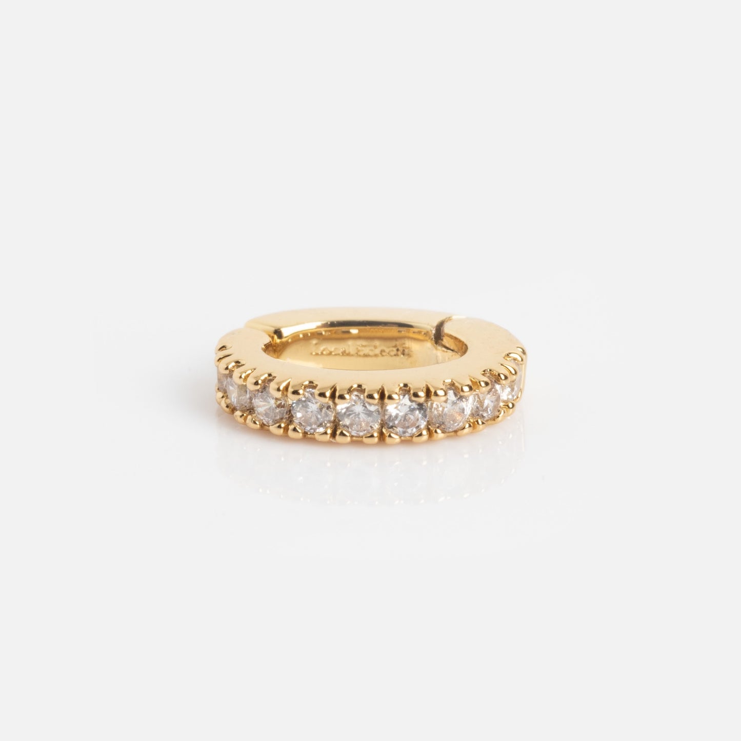 The Ring Clip, a small gold hoop earring, lies flat on a white surface and features sparkling clear gemstones set along its outer edge.