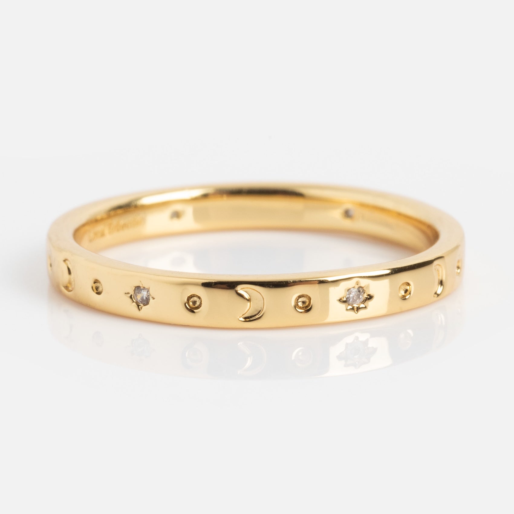 The Bold Moon Beam Stacking Ring is a thin gold band engraved with crescent moons, small circles, and star shapes, each accented by tiny gemstones spaced evenly around the ring.