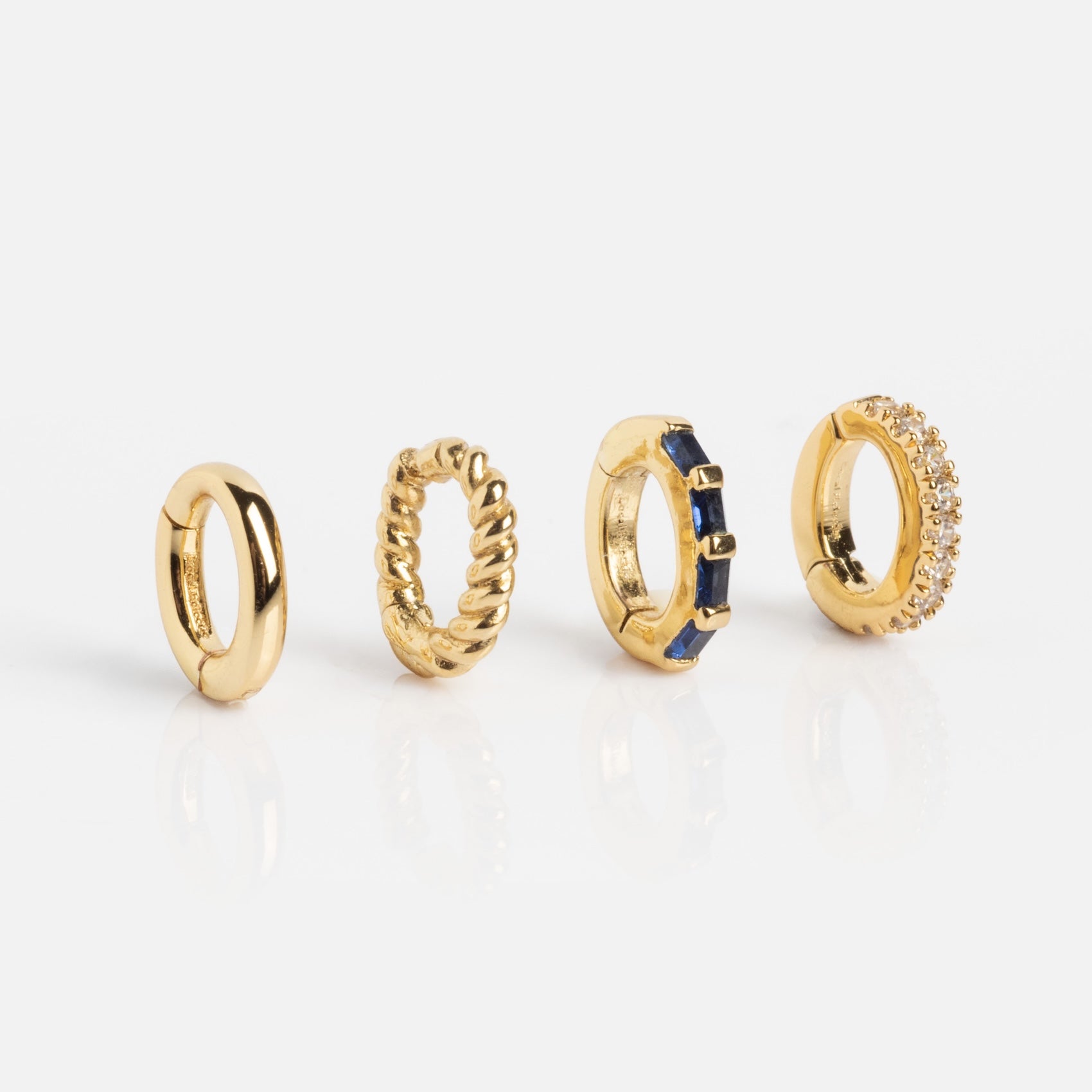 Four small gold Ring Clip earrings—plain, twisted rope, blue gemstone accents, and clear gemstones—each with a unique design, displayed upright in a row on a white background.