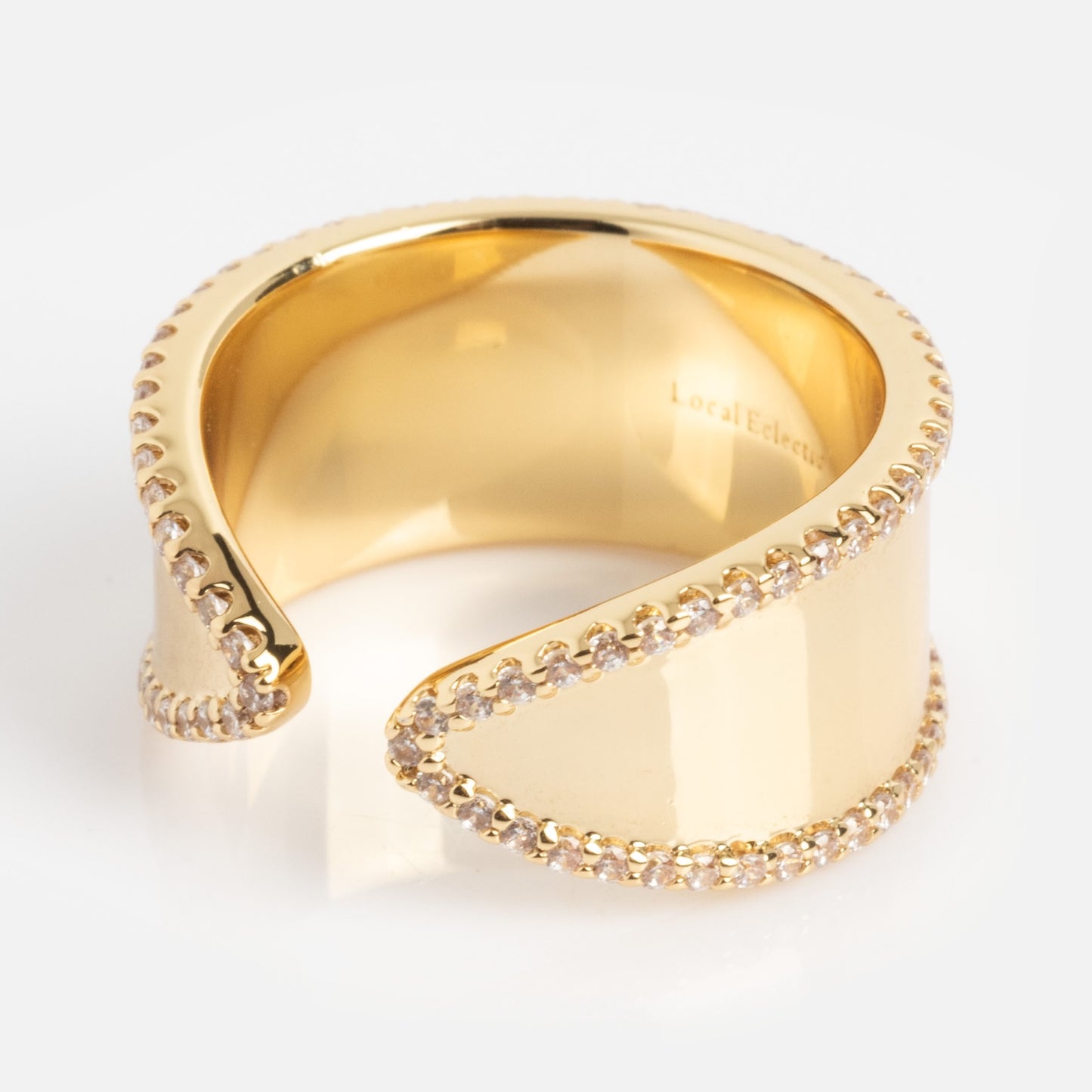 The Open Front Cigar Pinky Ring is a wide, gold-toned ring with an open-ended, wavy design and edges embellished with small, sparkling clear stones.