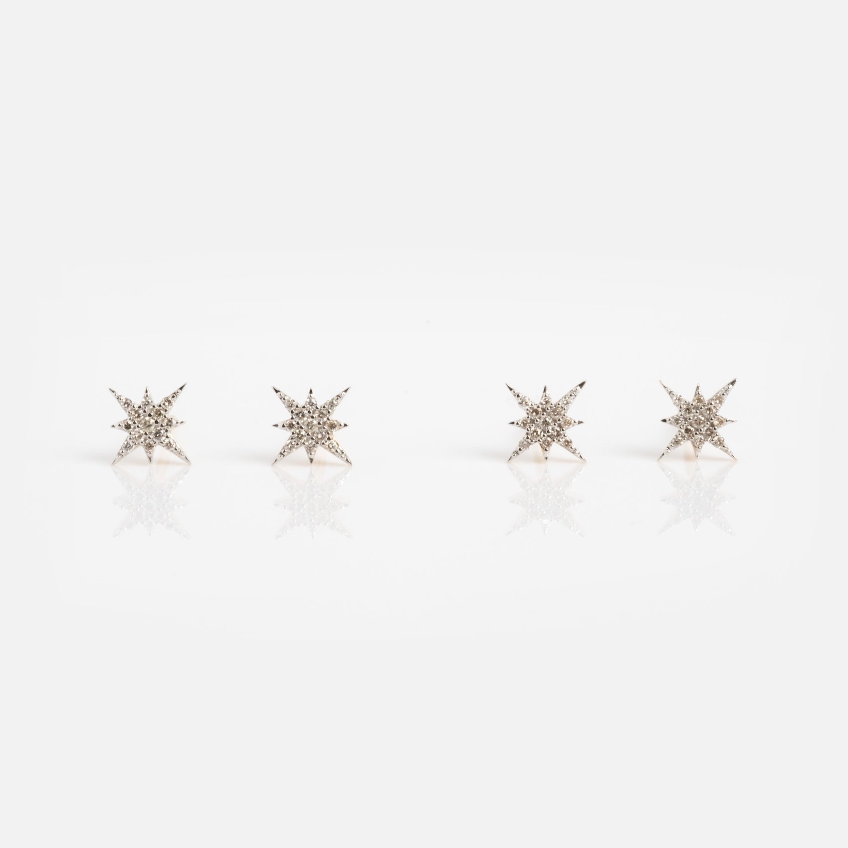 Four Solid Gold Diamond Starlight Studs, inspired by celestial designs and set with sparkling diamonds, are arranged in a row on a white background with a subtle reflection beneath them.