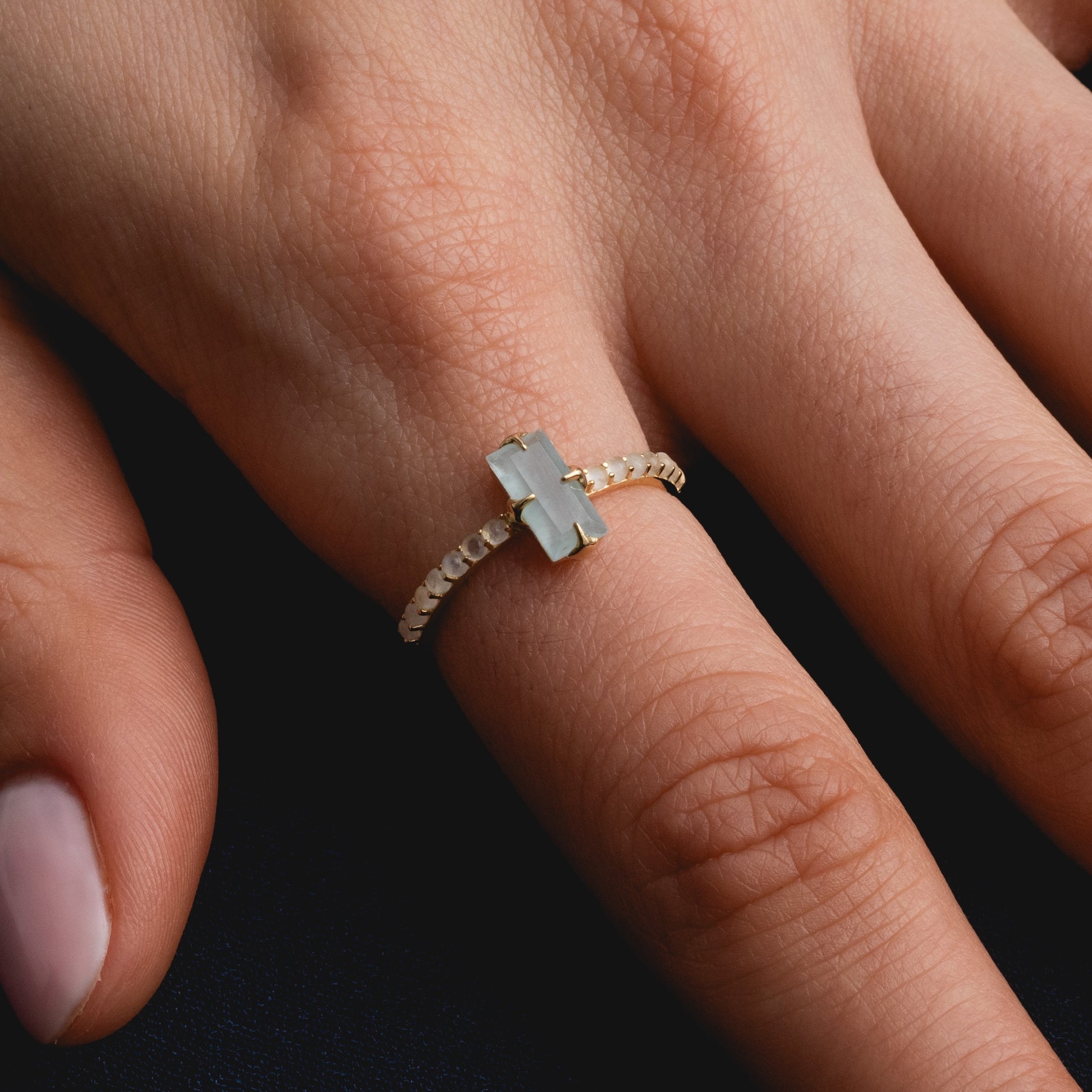 A close-up of a hand wearing the Solid Gold 2026 March Capsule Delicate Aquamarine Solitaire Ring, featuring a rectangular light aquamarine stone set horizontally on a textured gold band, resting on a dark surface.