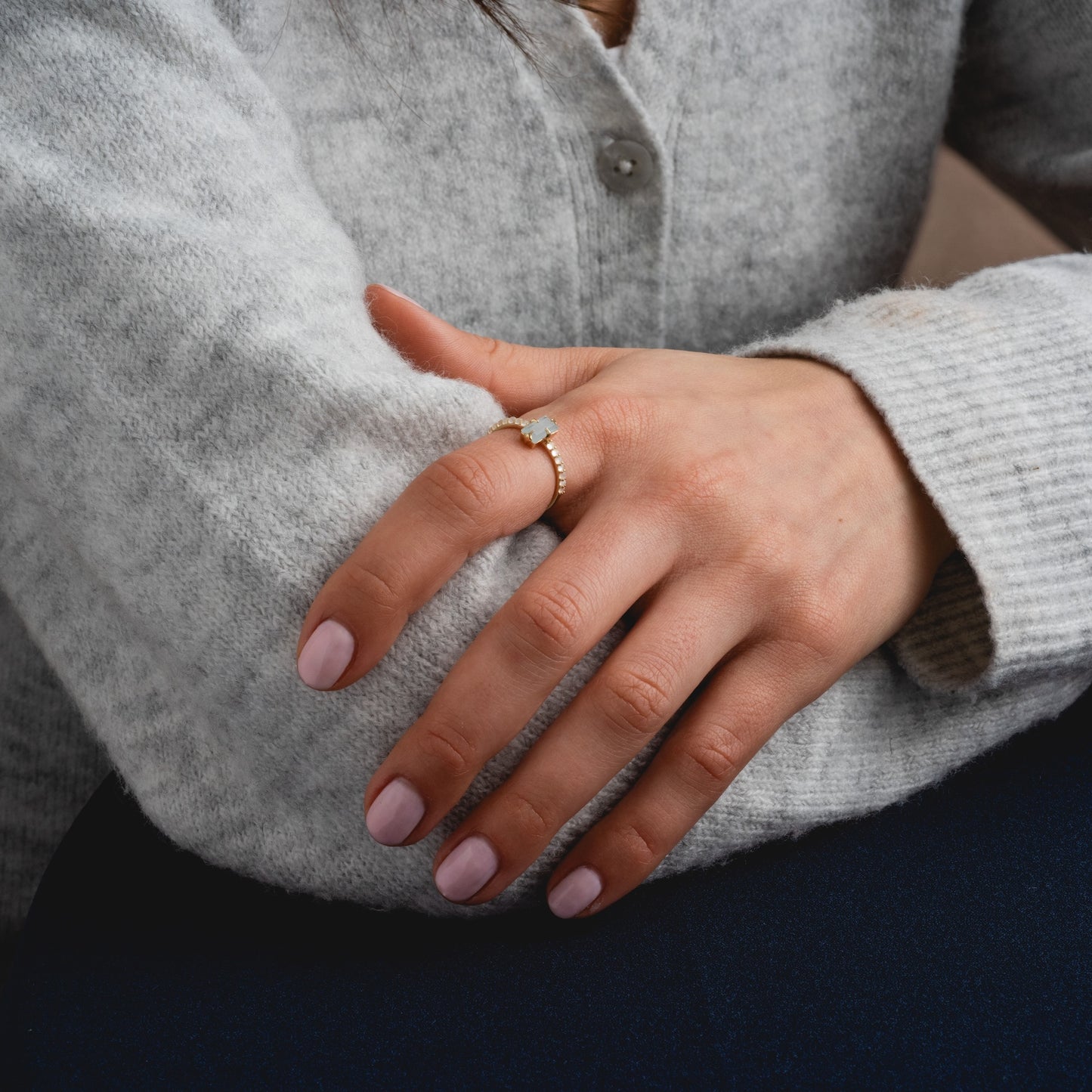 A person wearing a soft, light gray sweater crosses their arms, showcasing the Solid Gold 2026 March Capsule Delicate Aquamarine Solitaire Ring on their finger. Their neatly manicured nails are painted pale pink.