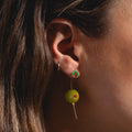 Close-up of a person’s ear wearing two trendy earrings: a small gold hoop and Olive Drop Earrings—shaped like green olives with red pimento, gold toothpick, and green leaf detail.