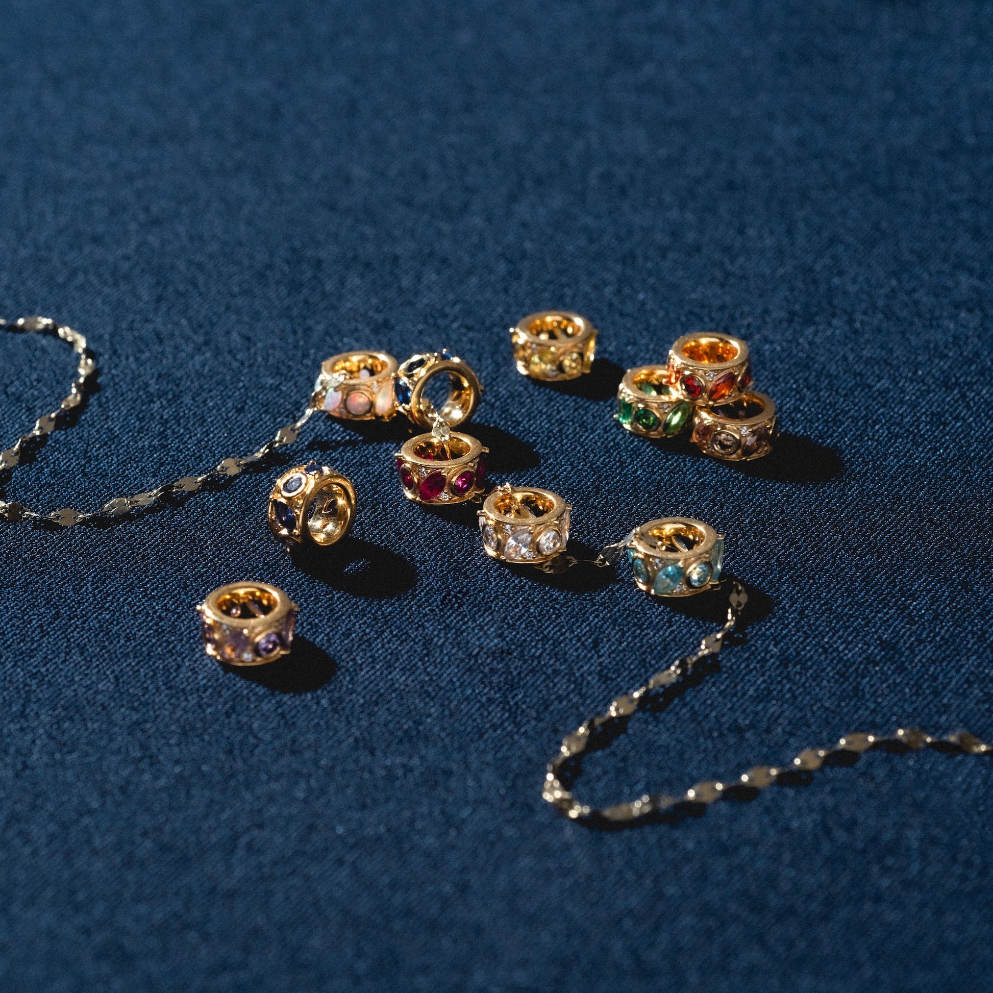 The Shades of You Birthstone Spacer Charm is displayed on dark blue fabric beside two delicate gold chains, creating a vibrant and elegant jewelry arrangement.