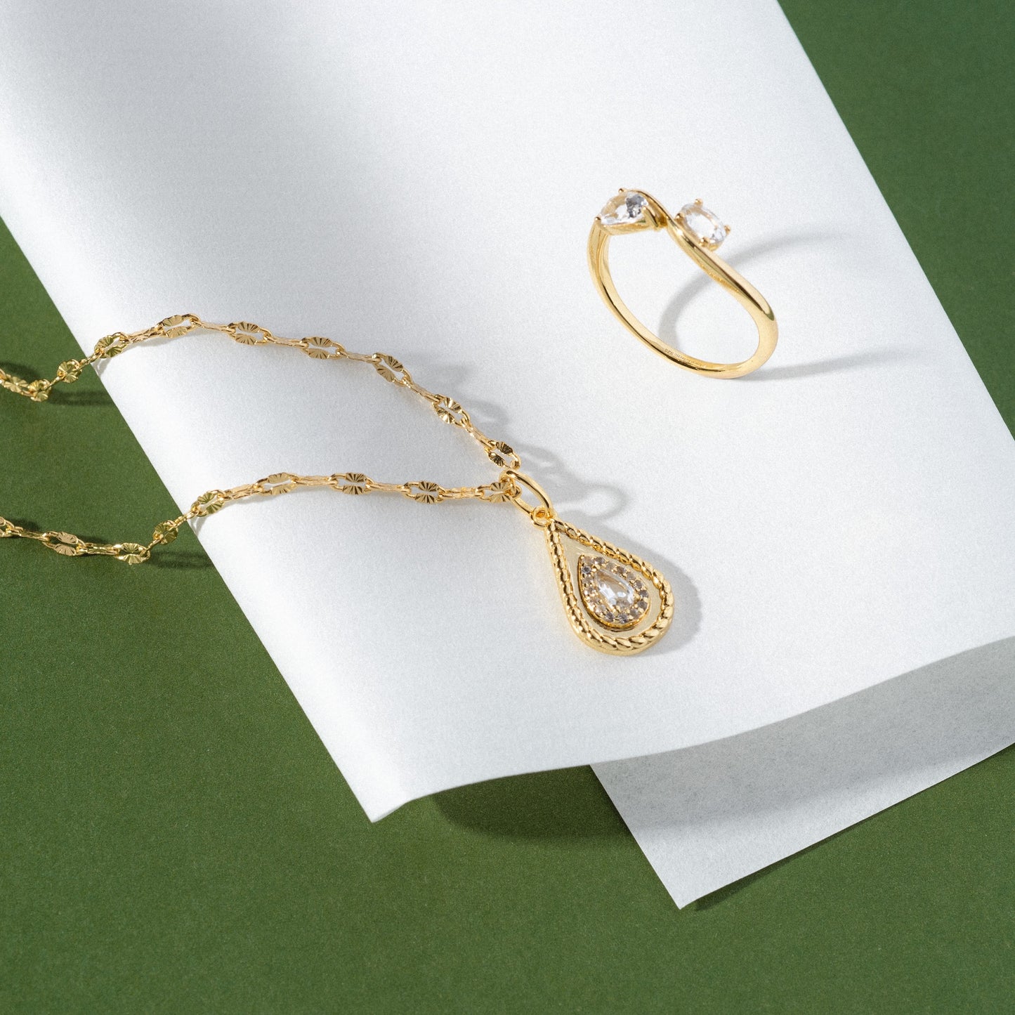 The 2026 April Capsule Tethered Teardrop White Topaz Charm, a yellow gold vermeil necklace with a white topaz pendant, and a gold ring with two clear stones are displayed on white paper over a green surface.