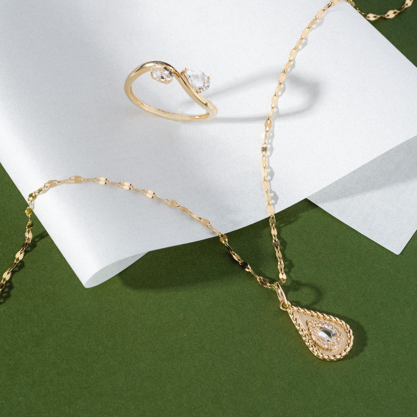 The Solid Gold 2026 April Capsule Tethered Teardrop White Topaz Charm is displayed with a gold ring set with a clear gemstone on white paper against a green background.