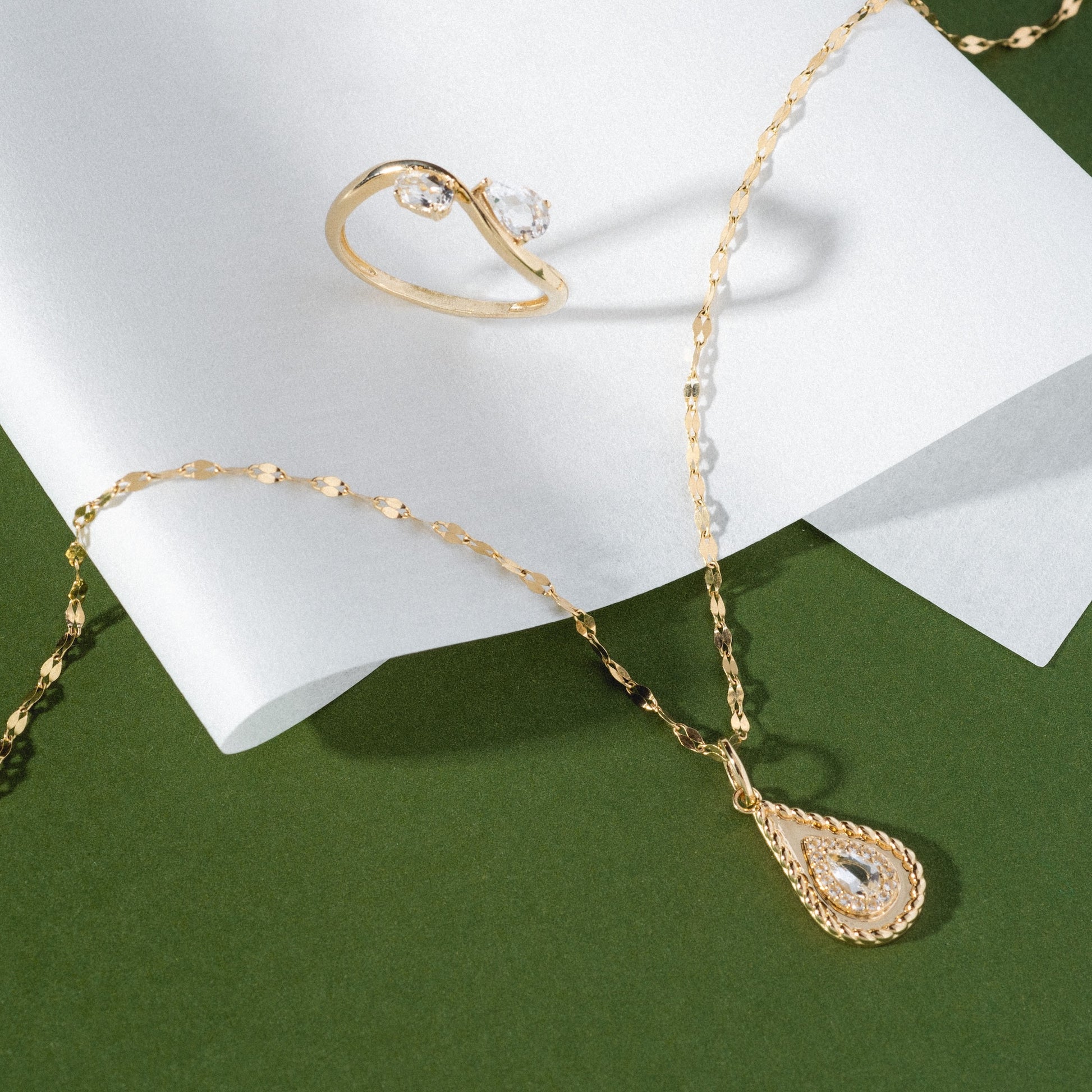 The Solid Gold 2026 April Capsule Tethered Teardrop White Topaz Charm is displayed with a gold ring set with a clear gemstone on white paper against a green background.