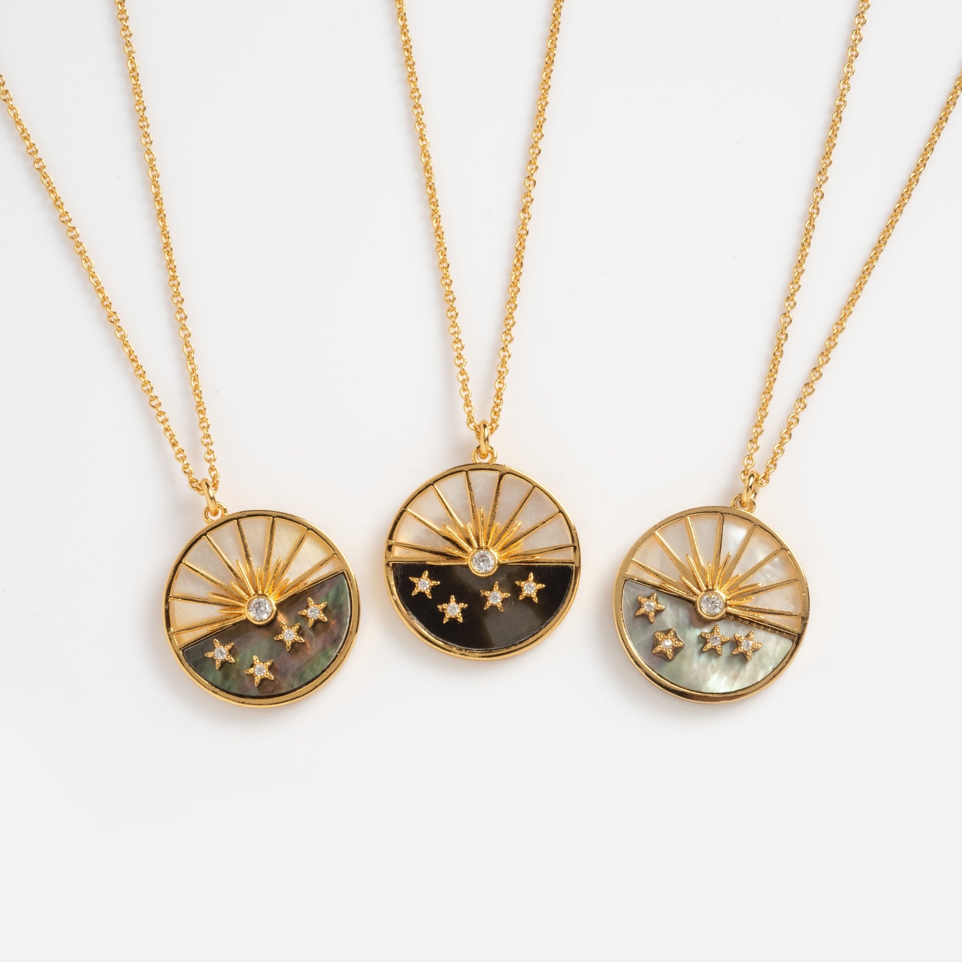 The Abalone and Mother of Pearl Horizon Necklace features a round sunburst pendant with small stars and gemstones at the center, accented with black, white, or green abalone sections, all displayed on gold chains against a white background.