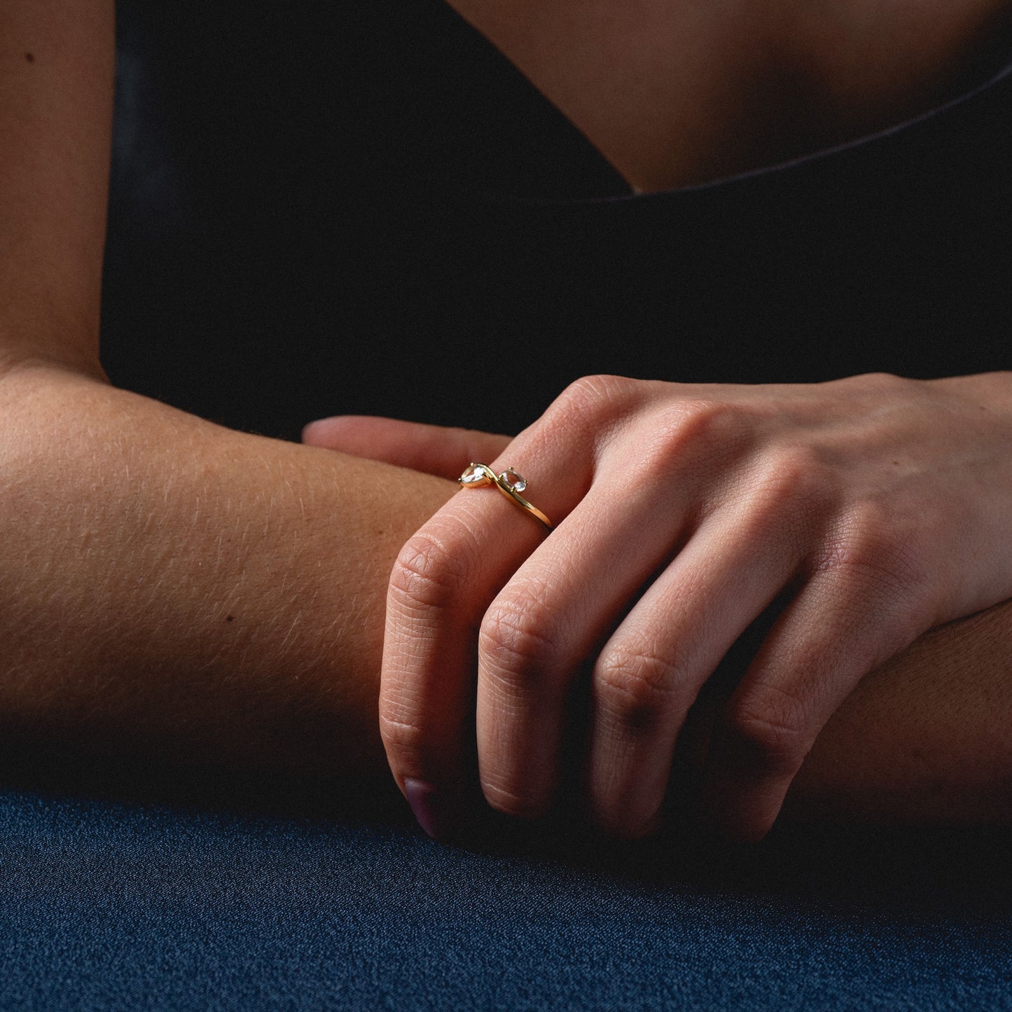 A close-up of a hand resting on dark blue, wearing the 2026 April Capsule Floating Duo White Topaz Ring in yellow gold vermeil—an elegant April birthstone piece. The person wears a sleeveless dark top.