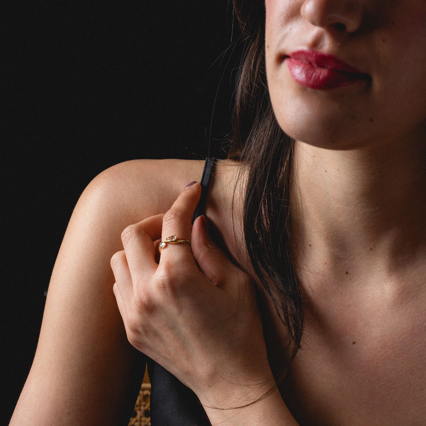 A woman with long dark hair and red lipstick touches her shoulder strap while wearing the 2026 April Capsule Floating Duo White Topaz Ring in yellow gold vermeil, against a dark background. She is dressed in a black top.