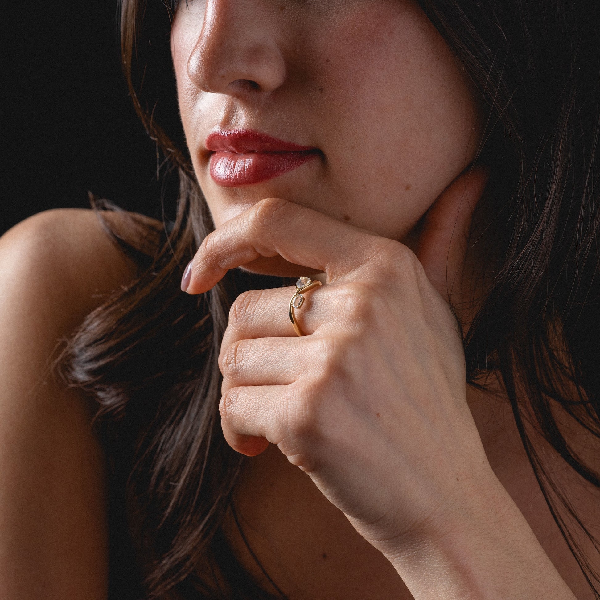 A woman with long dark hair and red lipstick rests her chin on her hand, showcasing the Solid Gold 2026 April Capsule Floating Duo White Topaz Ring. The dark background highlights her thoughtful look and the exquisite limited edition jewelry piece.