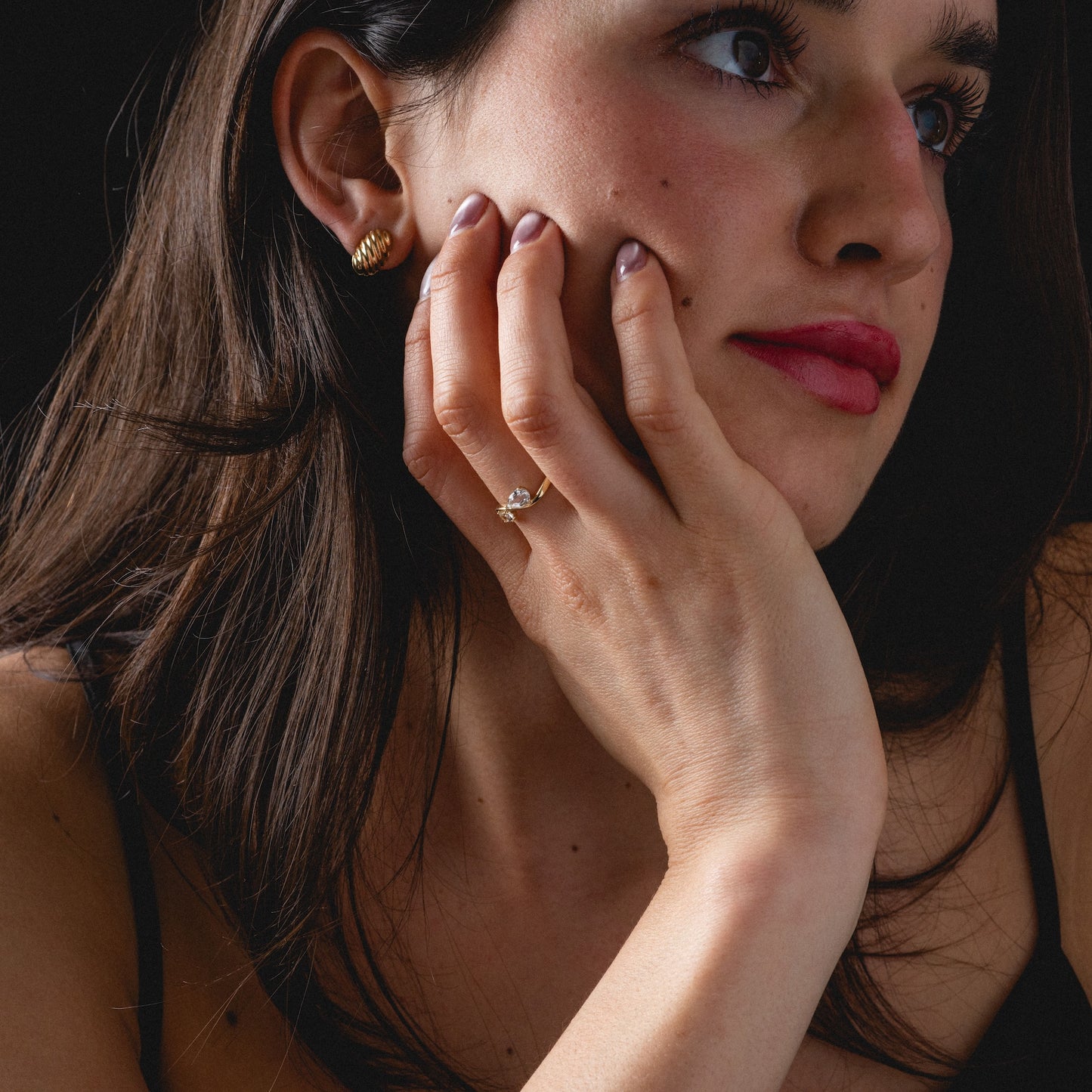 A woman with long dark hair rests her chin on her hand, wearing the limited edition Solid Gold 2026 April Capsule Floating Duo White Topaz Ring and gold earrings. She gazes thoughtfully to the side, in subtle makeup and a black top against a dark background.