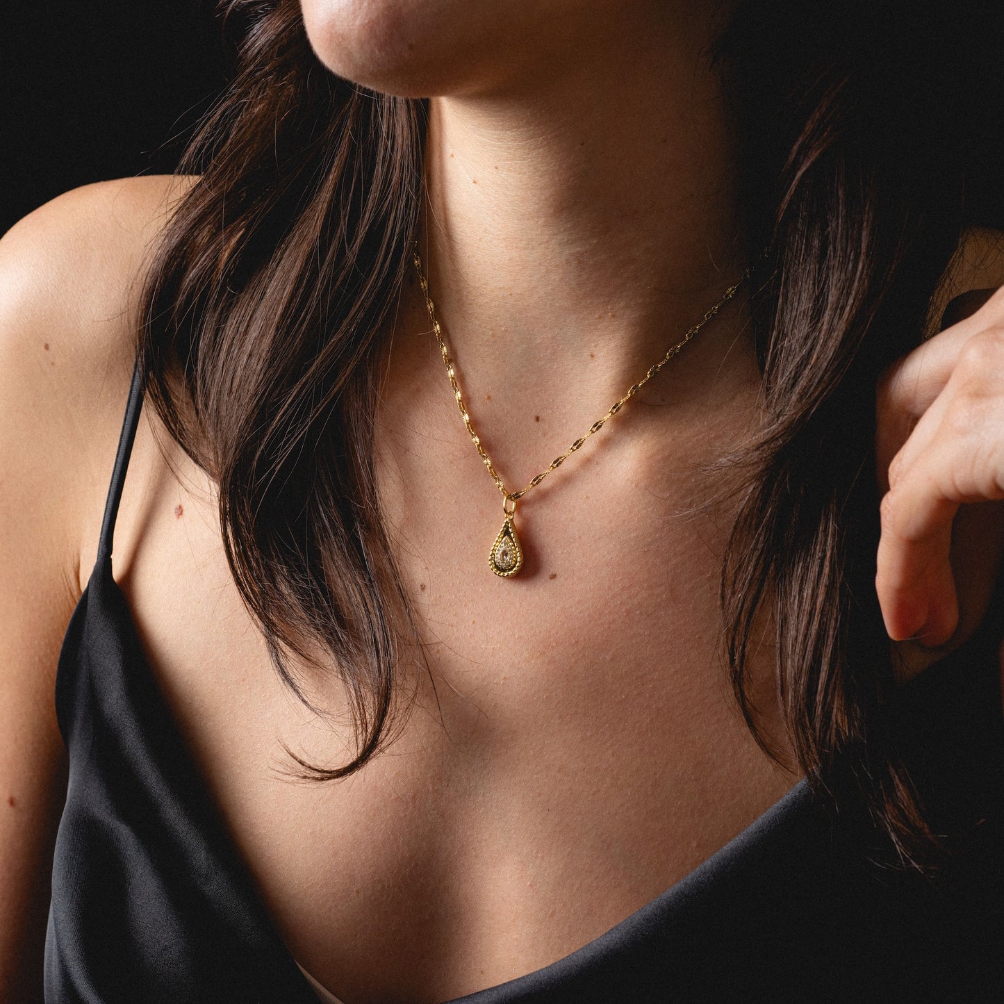 A woman in a black spaghetti strap top showcases the 2026 April Capsule Tethered Teardrop White Topaz Charm, a yellow gold vermeil necklace with a teardrop-shaped white topaz pendant. Her dark hair cascades over her shoulders against a dark backdrop.