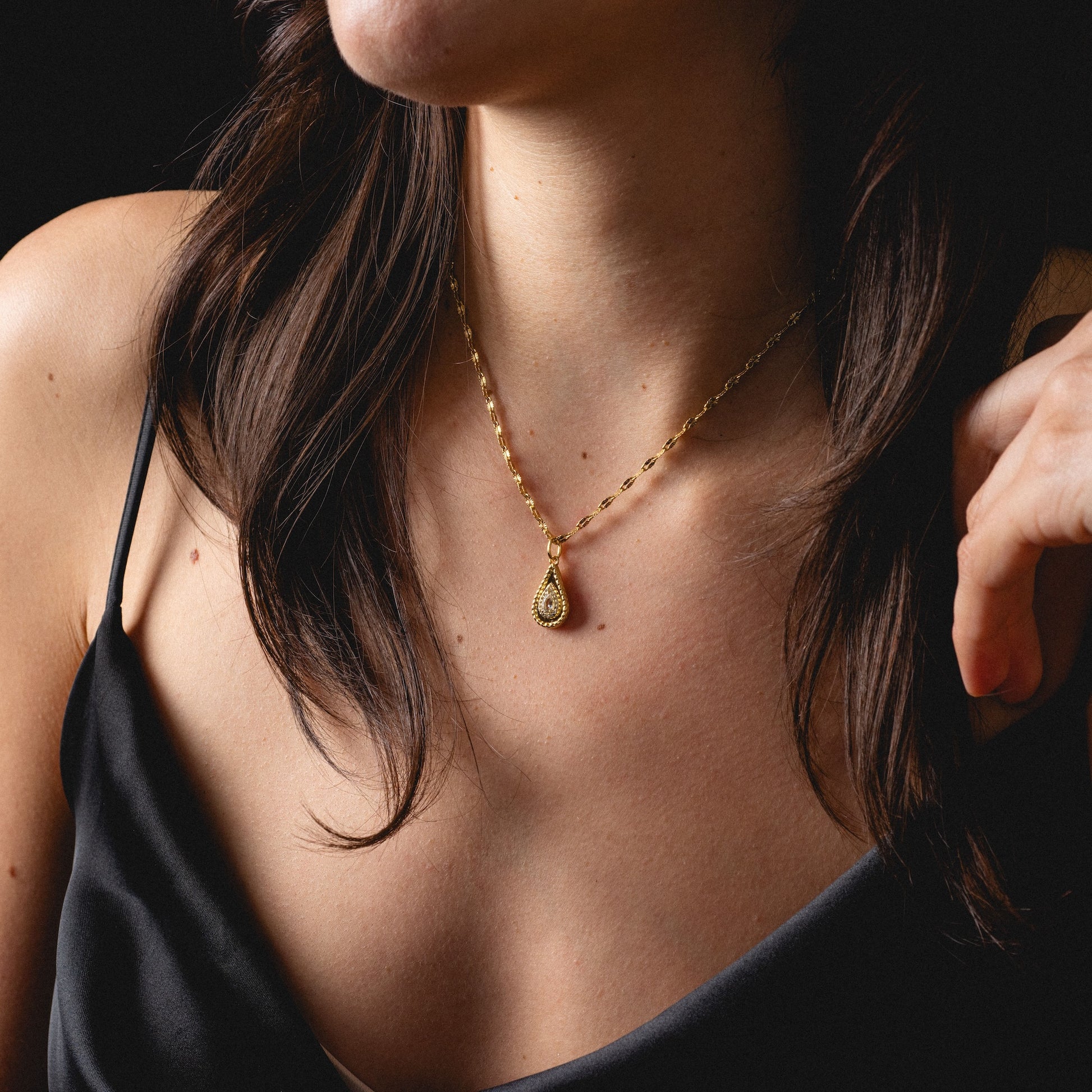 A woman in a black spaghetti strap top showcases the 2026 April Capsule Tethered Teardrop White Topaz Charm, a yellow gold vermeil necklace with a teardrop-shaped white topaz pendant. Her dark hair cascades over her shoulders against a dark backdrop.