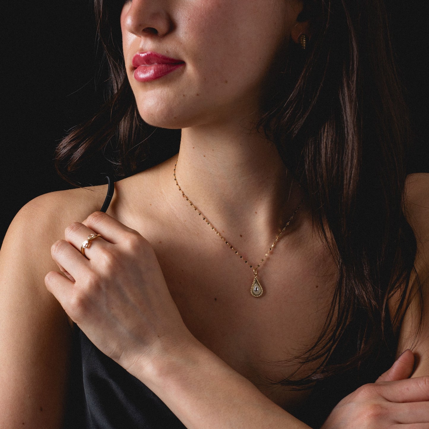 A woman in a black spaghetti strap top models the Solid Gold 2026 April White Topaz Capsule Set, featuring a teardrop pendant necklace and gold ring. She has loose dark hair, pink lips, and poses against a black background.
