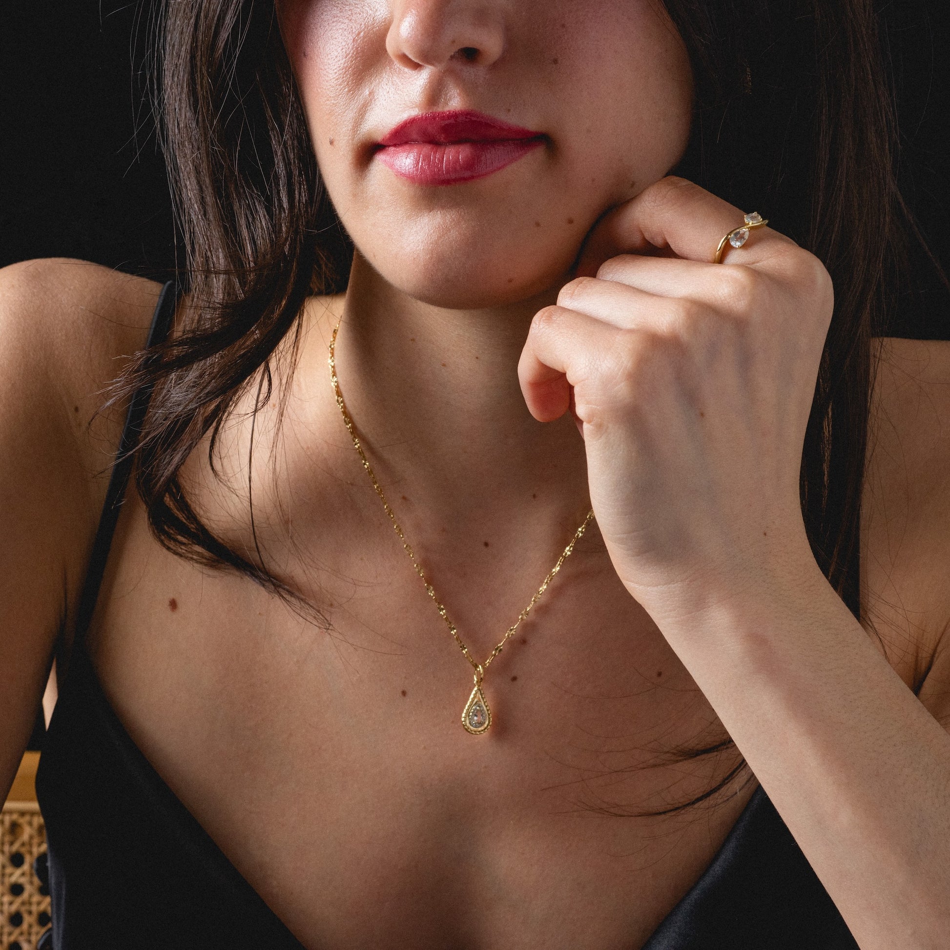 A woman in a black top models the 2026 April White Topaz Capsule Set with Free Gift, featuring a gold vermeil pendant and white topaz ring. She has dark hair, deep red lips, and rests her chin on her hand.