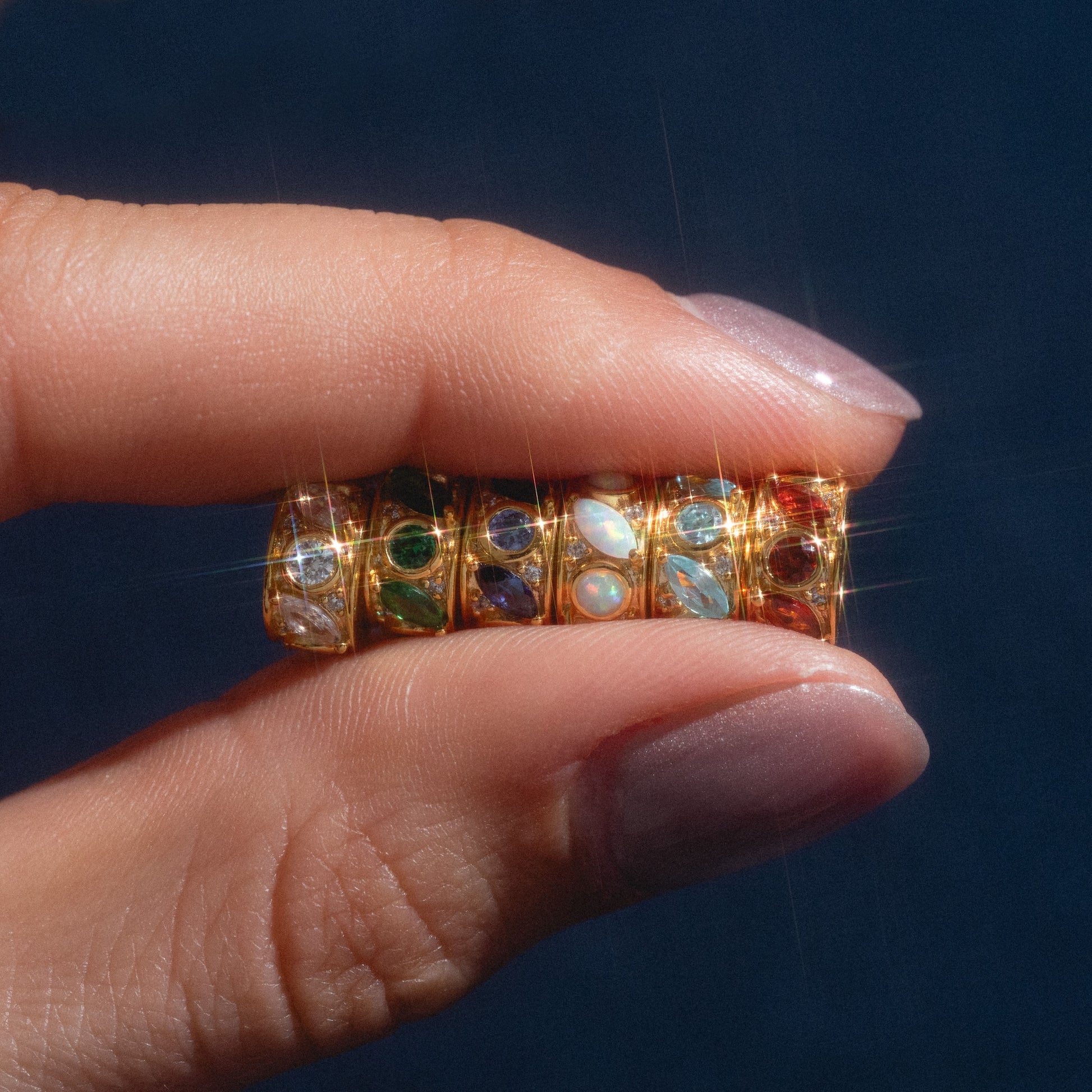 A manicured hand holds four Shades of You Birthstone Spacer Charms—gold rings set with colorful gemstones—against a dark background.