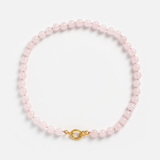 The Beaded Gemstone Charm Holder Necklace features round, pale pink beads and a gold clasp, displayed in a circular shape on a plain white background—a delicate statement piece for gemstone jewelry lovers.