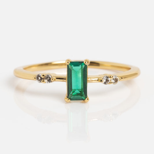 The 2026 May Capsule Delicate Emerald Solitaire Ring showcases a rectangular emerald centerpiece accented by two small round diamonds on each side, elegantly displayed against a white backdrop.