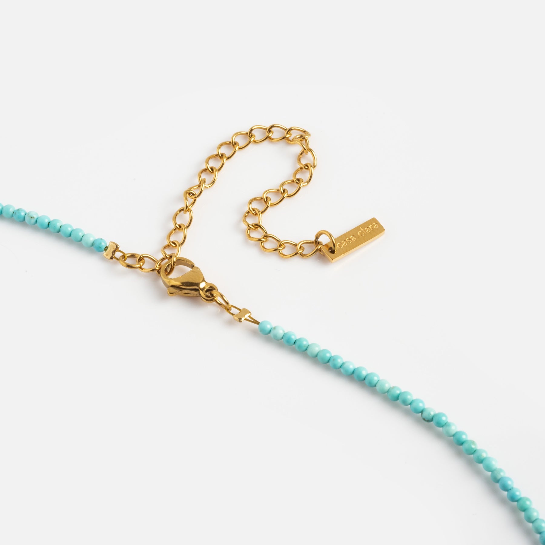 A close-up of the Byron Necklace, featuring small turquoise beads, a gold lobster clasp, and an adjustable gold chain with a rectangular engraved tag on a white background.