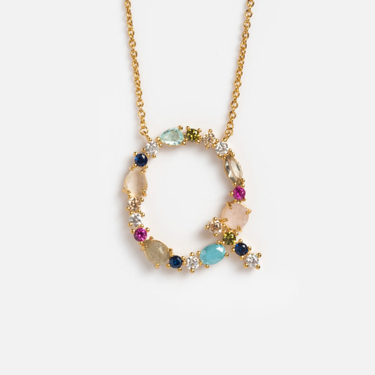 The Colorful Monogram Pendant is a gold necklace with a circular pendant, accented by vibrant gemstones and sparkling cubic zirconia crystals arranged asymmetrically, shown on a plain white background.