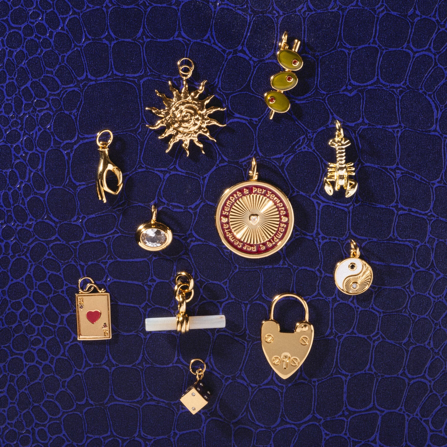 Twelve Charms by Mod + Jo, including a sun, olive branch, skeleton, yin-yang, lock, playing card, dice (a classic good luck charm), and compass, are displayed on a dark blue faux-crocodile background.
