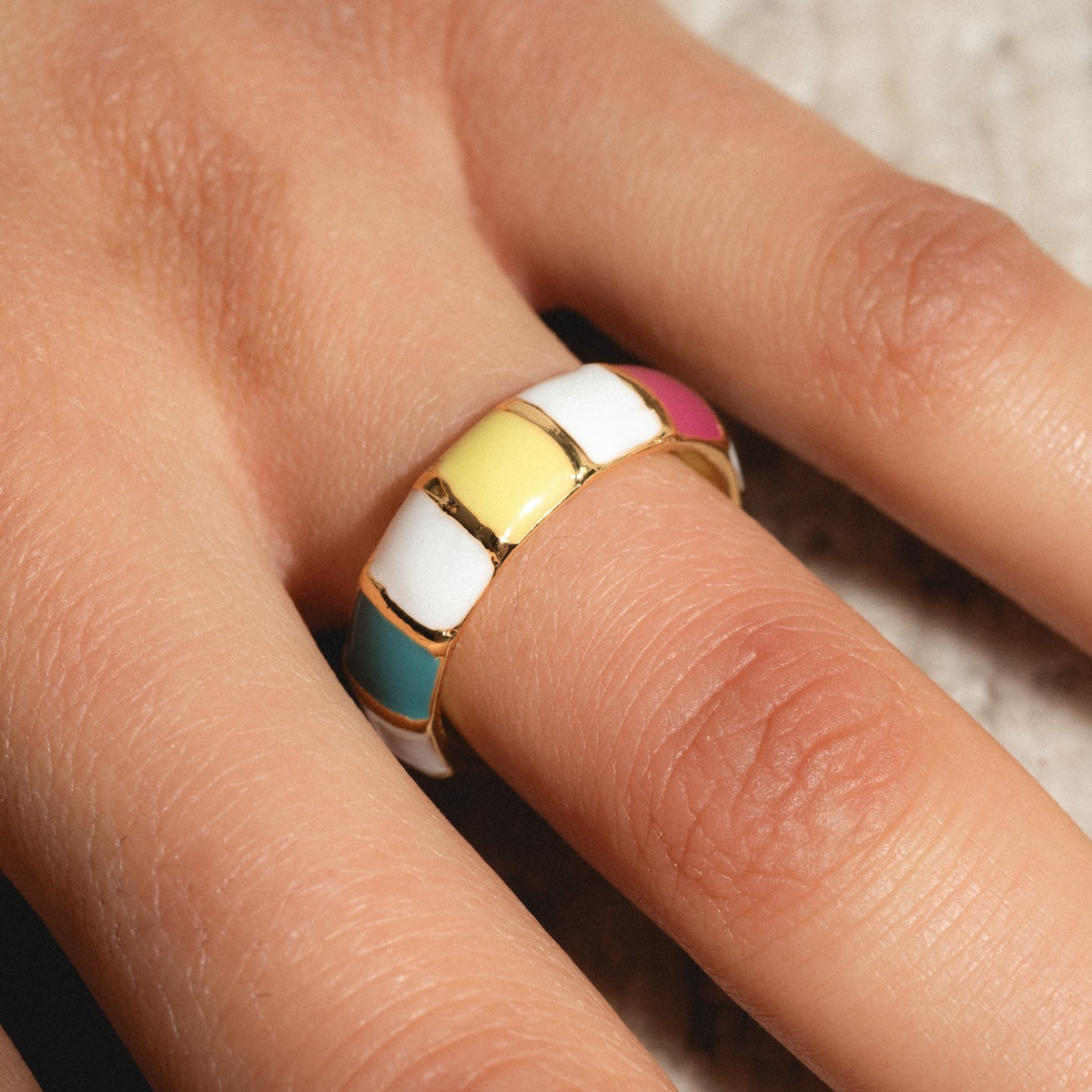 A hand wears the Chroma Ring—24k gold plated with colorful enamel sections in white, yellow, teal, and pink.