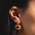 Close-up of a womans ear wearing Zand Ruby Drop Earrings—gold statement earrings with a round stud and an oval-shaped red gemstone, reminiscent of ruby drops, framed in decorative gold.
