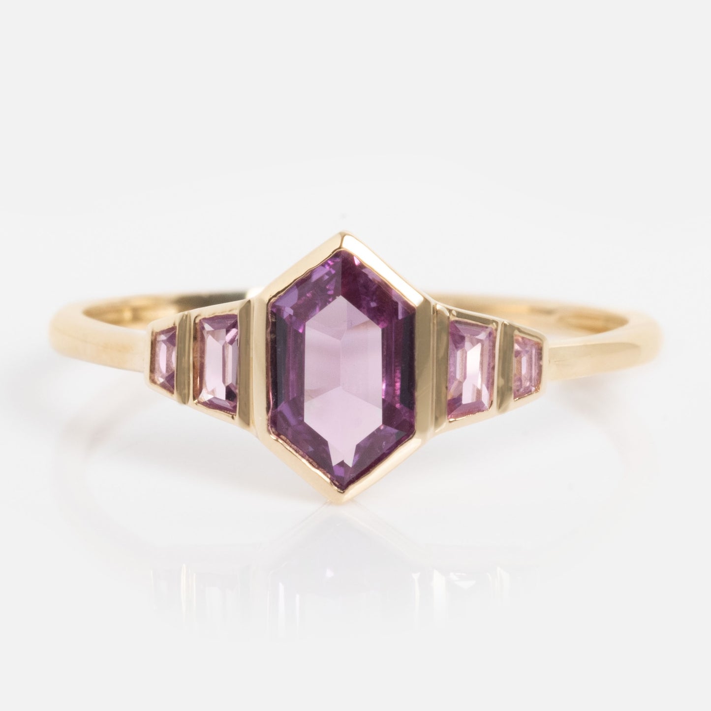 The Solid Gold 2026 June Capsule Art Deco Alexandrite Ring showcases a central hexagonal purple alexandrite, flanked by two smaller rectangular stones on each side, all set on a sleek gold band against a white background.