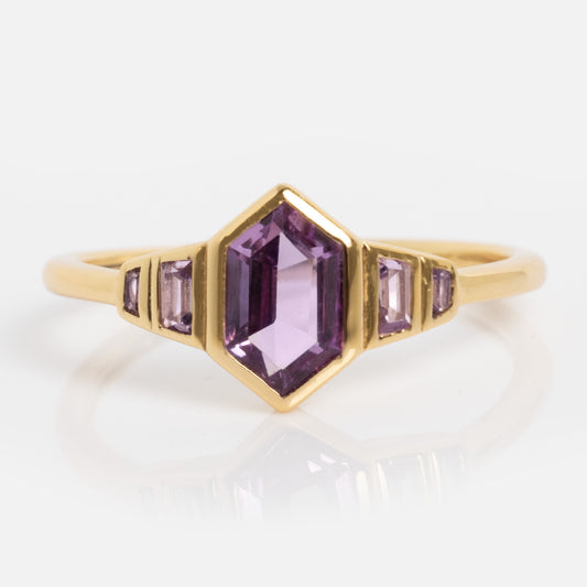 The 2026 June Capsule Art Deco Alexandrite Ring features a gold band with a central hexagonal purple gemstone and two smaller rectangular purple stones on each side, displayed on a plain white background.