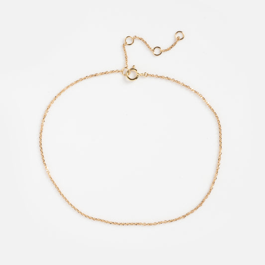The Solid Gold Simple Glimmer Gold Chain Anklet features a delicate gold chain with a small clasp and adjustable links, displayed in a loose oval on a plain white background.