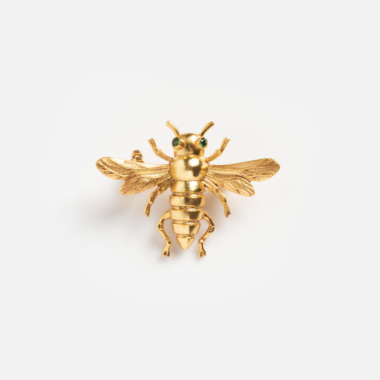 The Queen Bee Brooch features a gold bee design with intricate wings and green gemstone eyes, showcased on a plain white background.