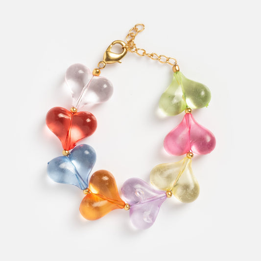 The Glossy Hearts Bracelet features heart-shaped glass beads in clear, red, blue, orange, purple, yellow, pink, and green, linked by small gold beads and finished with a gold chain clasp. Displayed on a white background.