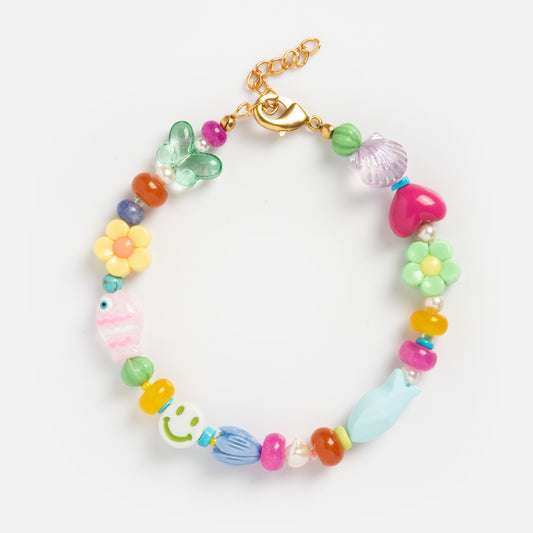 The Whimsy Medley Beaded Anklet features colorful beads and assorted charms— butterfly, fish, flower, heart, and smiley face— with a gold clasp and adjustable chain, shown on a white background.