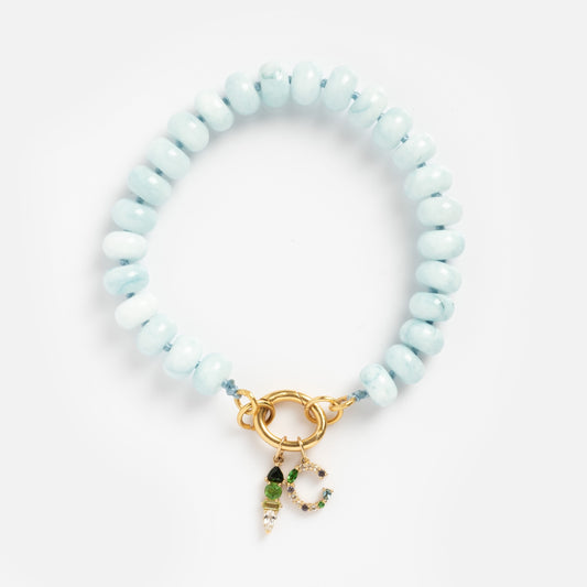 The Beaded Gemstone Charm Holder Bracelet features light blue round beads, a gold clasp, and two small gold and green charms, elegantly displayed on a white background.
