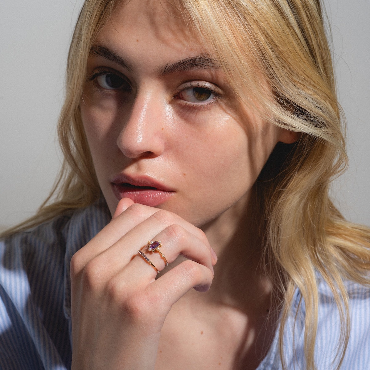 A young woman with blonde hair and light skin gazes thoughtfully at the camera, wearing a blue striped shirt and the Solid Gold 2025 June Capsule Alexandrite and Pearl Ring Set—stylish June birthstone jewelry. Free gift included.
