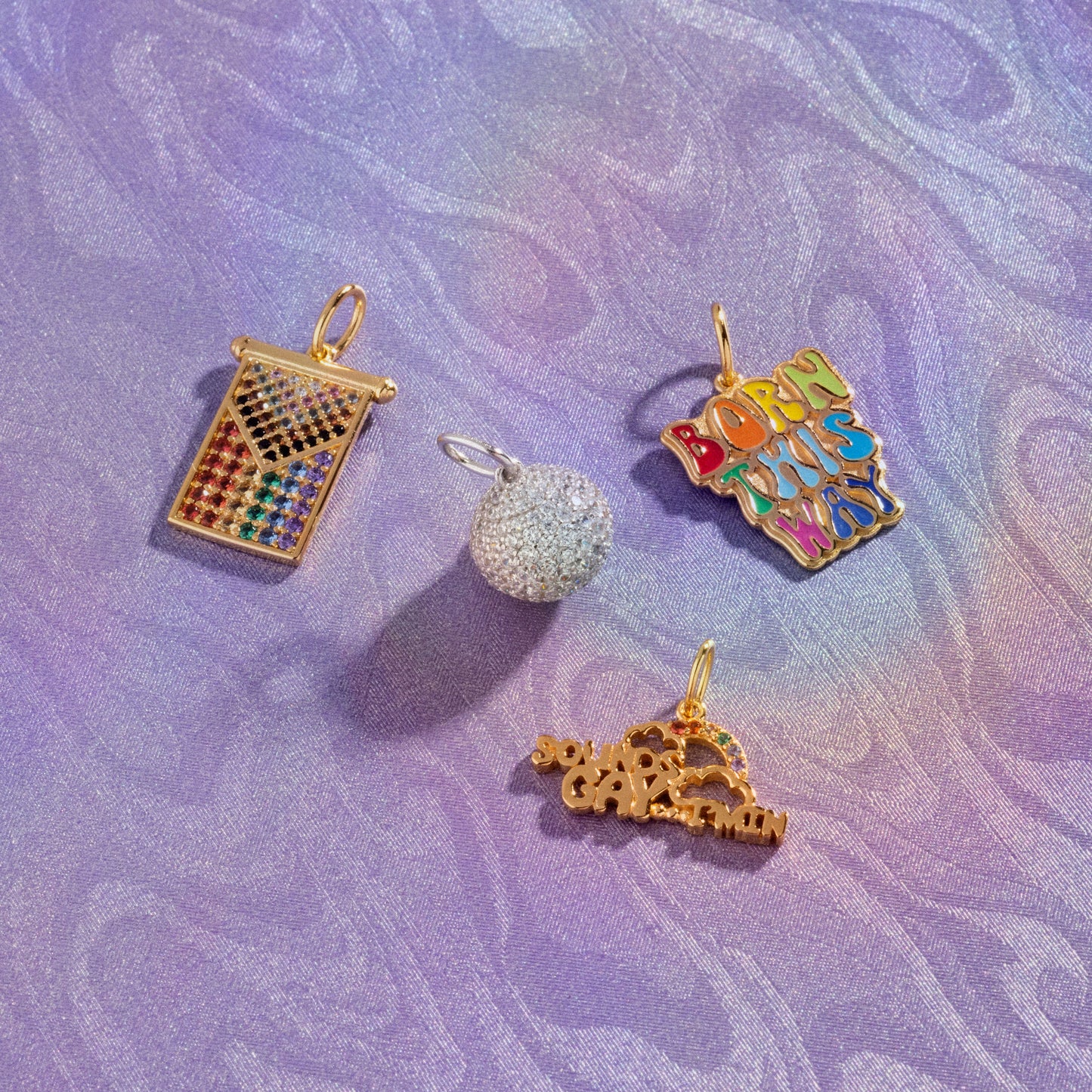 Limited Edition PRIDE Month Charms: four colorful pendants on a purple iridescent background—a rainbow gem grid, sparkly silver ball, BORN THIS WAY in rainbow letters, and a gold charm with “GAGA” and a heart.